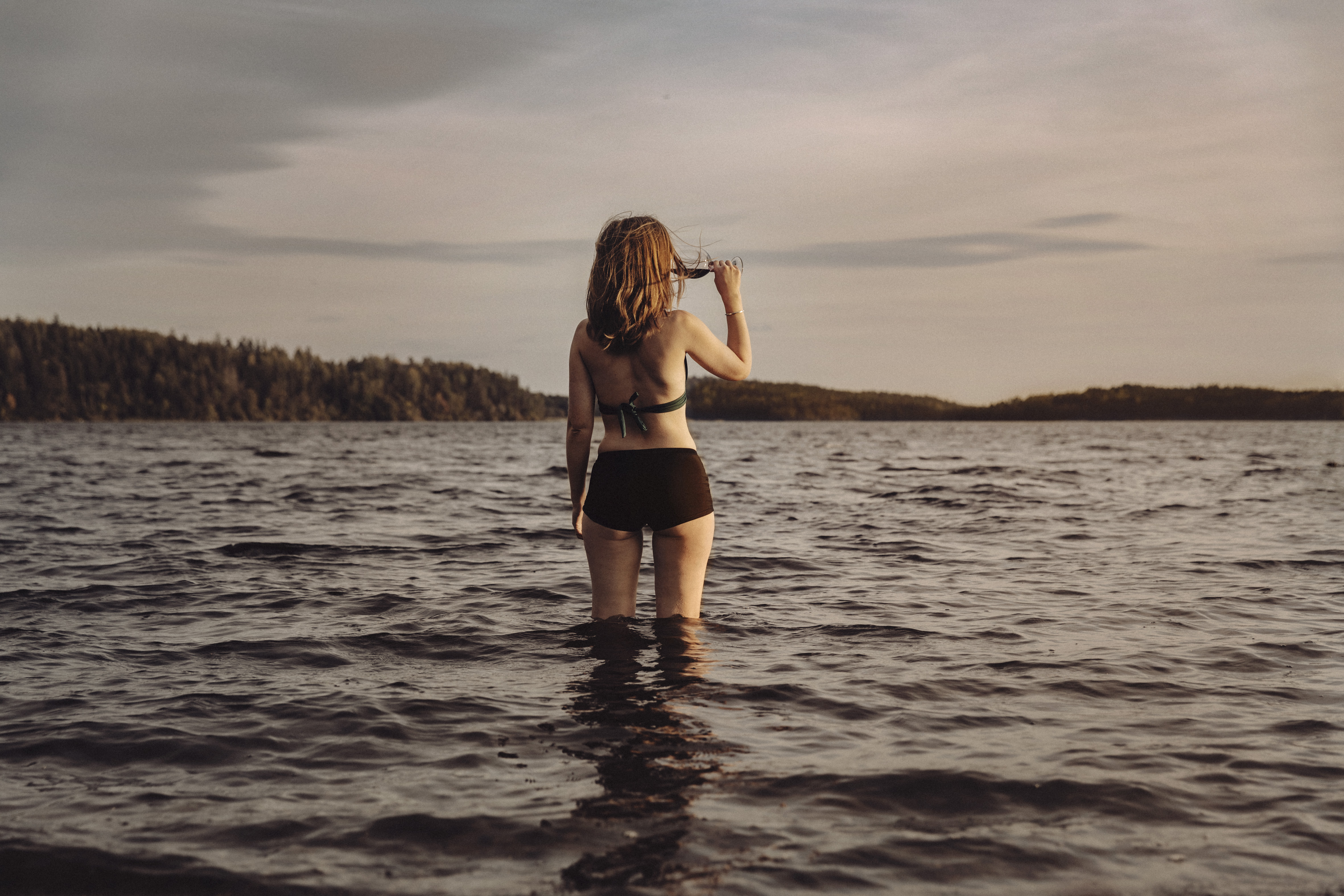 Person står i bikini med vatten upp till knäna och njuter av ett glas rödvin på Friibergh